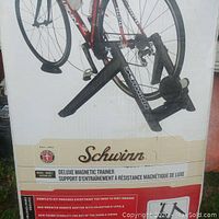 Bike mounted on Schwinn Deluxe Magnetic Trainer displayed on original box, showing black aluminum trainer frame with Schwinn logo, rear wheel holder, magnetic resistance mechanism, and adjustable remote shifter cable.