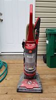 Full front view of red Hoover upright vacuum cleaner with attached hose and brush, placed on wooden deck outside white door.