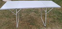 Fully extended folding utility table on grass, showing white surface and metal legs setup.