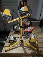 Photo shows yellow tripod supporting four work lights with wire cages, large Cyclops spotlight, and smaller work lights on wooden surface.