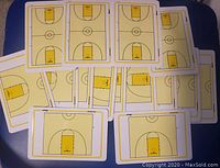 Photo of 15 pocket-sized basketball tactic boards laid overlapping on a dark surface, each showing a half-court diagram in yellow and white.