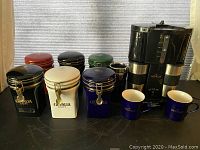 Image showing Gevalia coffee maker, six ceramic coffee canisters in red, black, white, green, and blue, two coffee cups with saucers, and a stainless steel insulated beaker.
