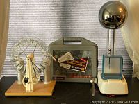 Wimshurst generator on left with large clear acrylic discs and metal components, microscope kit in center inside gray plastic case labeled Deluxe Microscope Set, and Van de Graaff generator on right with large metal sphere and white base.