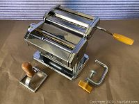 Pasta machine with removable crank handle and securing clamp laid out on surface.