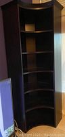 Tall corner shelf unit with dark wood finish, concave curved front, and four internal shelves, shown standing beside a wall and television.