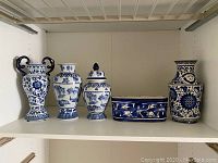 Four blue and white ceramic pieces displayed on a shelf: two vases, one urn with two handles, and one rectangular planter.