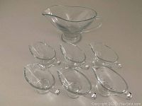 One large clear glass sauce jug with a wide oval body, handle, spout and pedestal base. Six smaller matching sauce jugs arranged in front, clear glass with handles and spouts.