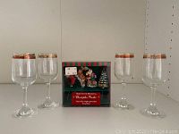 Set of four clear wine glasses with gold and rose decorative bands around their rim alongside boxed Christmas butter/cheese spreaders.