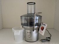 Front view of Breville Juice Fountain juicer including the main juicer body, clear plastic pulp container, and transparent measuring cup with markings. The juicer is silver with black accents and has a power cord attached.
