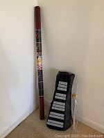 Full view of the didjerido standing upright next to the folded glockenspiel in case with mallets leaning against the wall.