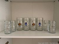 Wide view of all 8 dimpled glass beer mugs arranged in a row on shelf, showing two large Hofbrauhaus Munchen mugs in the center and smaller mugs on both sides.