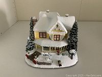 Front and side view of illuminated sculpture showing detailed snow-covered Victorian-style house with front porch, trees, lamp posts, and small figures.