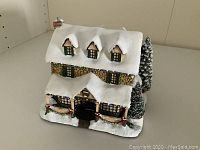 Front angled view of the illuminated ceramic cottage sculpture with snow and holiday decorations.