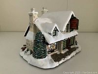 Side angle of Thomas Kinkade Santa's Workshop Toys illuminated village house showing snow-covered roof and Christmas tree.