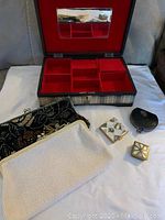 Photo showing opened plaid jewelry box with red velvet compartments and mirror inside lid, positioned behind purses and compact cases laid out on white surface