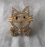Front view of gold-tone cat brooch with clear rhinestones, black eyes, gold whiskers, and rhinestone bow tie on white cloth background.