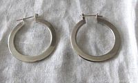 Pair of flat, polished sterling silver hoop earrings lying on light fabric background.