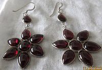 Pair of flower-shaped sterling silver earrings with six teardrop garnet stones forming petals around a round garnet center, plus a smaller teardrop garnet above each flower, displayed on white cloth.
