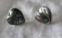 Front view of a pair of heart-shaped stud earrings with iridescent abalone shell in sterling silver setting on fabric background.