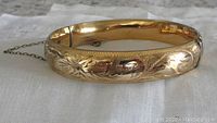 Close-up photo showing the gold filled hinged bangle with floral engravings and attached safety chain on a white linen background.