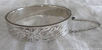 Silver hinged bangle bracelet with detailed floral engraving and safety chain visible, laid on fabric.