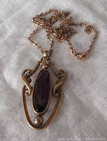 Front view of gilded brass pendant showing ornate scroll and bead frame around oval purple faceted glass stone on chain.