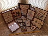 Overview of twelve hand cross-stitched pictures in various wooden frames arranged on beige carpet near window with blinds