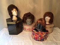 Four wigs placed on three decorative floral hat boxes and one black hexagonal hat box with chain detail arranged on white cloth surface.