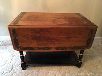 Full front view of the Hitchcock style drop leaf tea table closed, showing wood grain and painted decorative border on the top edges including floral motifs, supported by spindle legs and a black lower shelf.
