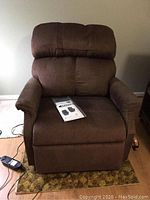 Front view of brown textured fabric power lift recliner showing seat, back, arms, wired remote and paperwork packet on seat.