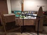 Photo showing wooden drying rack, black metal rolling garment rack, ironing board, and various laundry products on white tablecloth-covered table.