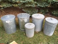 Five galvanized metal trash cans with lids, three large and two smaller, shown outdoors on grass with trees in the background.