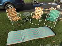 Three folding chairs in woven webbing (two orange/beige stripes, one green/white stripes), small green folding slatted table, and striped outdoor mat laid on grass.