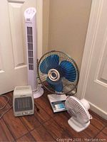 Four small appliances on floor: Maxi Heat heater, white tower fan, blue metal blade fan, small white desk fan.