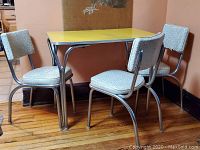 Photo showing 1950s style square table with yellow top and four chairs with white vinyl upholstery and chrome frames in corner setting.