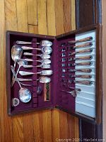 Full view of silverware set in original maroon velvet-lined case showing teaspoons, forks, and serving utensils arranged neatly.