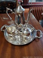 Full tea set on table showing coffee pot, sugar bowl, creamer, and platter with engraved surface and handles.