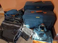 Photo showing the full lot of luggage pieces arranged on a carpeted floor; includes large blue suitcases, black suitcase, and soft bags.