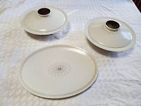 Two covered vegetable tureens and one oval serving platter displayed on white tablecloth. Tureens have fitted lids with round knob handles. All pieces in white ceramic with patterned border and starburst center design.