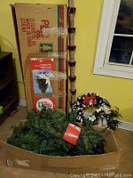 Boxed sections of artificial Christmas tree with loose branches and tree stand, wreaths and garland placed in front of box