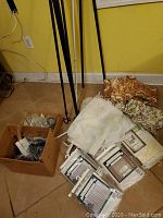 Photo showing boxed curtain hardware and folded vintage curtain fabrics, including floral and white fabric pieces, beside curtain rods on floor.