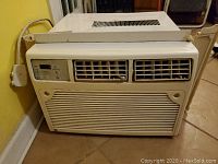 Front view of large GE window air conditioner unit placed against a yellow wall and brown floor, showing vents and overall condition.