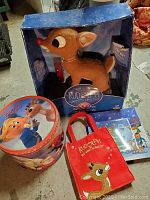 Newborn Rudolph plush-style action figure in blue original box showing wear, alongside popcorn tin, red tote bag, and holiday-themed tin box.
