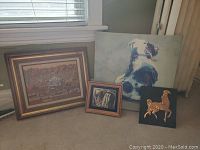 Four pieces of wall art including dog canvas, landscape painting, small figurative sketch, and horse plaque arranged on floor near window.