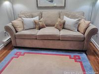 Front view of beige microfiber sofa bed with three seat cushions and eight pillows, placed on a colorful rug in a room with wood flooring.