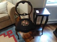 Vintage balloon back chair with dark wood frame and floral cross-stitch cushion with visible wear and cracks alongside a dark wood side table with lower shelf and wear from Bombay Co