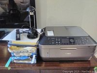 Canon Pixma MX870 printer on table next to paper reams and lamp cords, includes some paper packages and black desk lamp base.