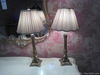 Two brass table lamps on white surface with pleated beige fabric lampshades and decorative bases, reflected in ornate wall mirror.