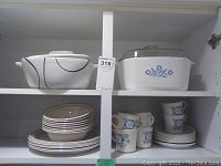 Shelf showing two casserole dishes on top shelf - one white with blue cornflower floral design and glass lid, other white with black swirl design with lid. Below are stacked plates, bowls, mugs, and saucers in blue cornflower pattern.