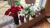 View showing several pots with faux flowers arranged on a windowsill, including red flowers, white flowers, and various pots of glass, ceramic, and plastic.